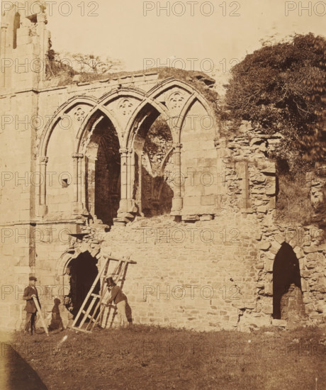 View of the refectory of Easby Abbey in Richmond, photograph, page, photographer: Joseph Cundall, Richmond (Yorkshire), photographer: Philip Henry Delamotte, c. 1851 - in or before 1856, Yorkshire, paper, albumen print, height 264 mm x width 221 mm, ruin of church, monastery, landscape with ruins (city(-scape) with figures, staffage), refectory, A large stone ruin with three pointed arch openings featuring decorative circular motifs sits against a grassy foreground, adjacent to a rough stone wall and a dense leafy tree on the right; two men wearing coats and hats stand near a wooden ladder leaned against the wall, one holding the ladder and the other holding a plank, while shadows fall on the masonry and grass.