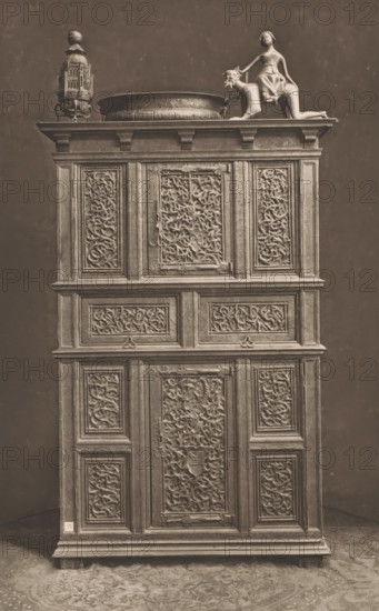 15th century German wooden cabinet, at the 1877 Exposition Rétrospective in Lyon, France, page, photomechanical print, inscription: 'ARMOIRE, ALLEMAGNE_FIN DU XVe SIÈCLE, COLLECTION DE Mr CHABRIÈRES-ARLES.', inscription: 'EXPOSITION RÉTROSPECTIVE DE LYON, PAR JB GIRAUD.', number: 'PL. I.', Lyon, cliché maker: Dujardin, printer (person): Imprimerie Eudes, Paris, 1877 - in or before 1880, photographer, printer (person), paper, photoaquatint, height 233 mm x width 147 mm, ornamental cupboard (wood and other phytogenic material), A tall wooden cabinet with multiple carved panels occupies the frame, topped by a decorative arrangement that includes an ornate metal lantern, a shallow round basin with embossed patterns, and a sculptural figure of a person seated on a stylized animal; the cabinet surface shows many rectangular framed panels filled with intricate scrolling foliage carvings and decorative moldings, metal hinges and latches are visible on some panels, the wood has a worn, aged app...