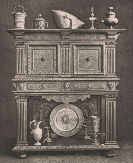 16th century French wooden sideboard, at the 1877 Exposition Rétrospective in Lyon, France, page, photomechanical print, inscription: 'CRÉDENCE, FRANCE_XVIe SIÈCLE, COLLECTION DE Mr CHABRIÈRES-ARLÈS.', number: 'PL. XXII.', inscription: 'EXPOSITION RÉTROSPECTIVE DE LYON, PAR JB GIRAUD.', Lyon, cliché maker: Dujardin, printer (person): Imprimerie Eudes, Paris, 1877 - in or before 1880, photographer, printer (person), paper, photoaquatint, height 227 mm x width 182 mm, ornamental cupboard (wood and other phytogenic material), A tall ornate wooden cabinet with carved panels and fluted columns displays a variety of metal objects on its top and lower shelf; the top surface holds a handled tankard, a domed container with side handles, a pointed helmet, a pedestal with a finial, and a bulbous vessel with a spout, all rendered in a tarnished metallic tone, while the lower shelf features a large decorative circular platter leaning against the back, a lidded ewer with a curved handle, two candles...