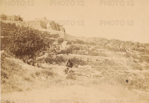 Bethlehem, with the church of nativity, Unknown man in a landscape with the Church of the Nativity in Bethlehem in the background, page, This photo is part of a cover with 7 sheets of albumen prints., photographer: Francis Frith, Jerusalem, 1857, paper, albumen print, height 158 mm x width 225 mm, Person, historical persons, church (exterior), city-view, and landscape with man-made constructions, Palestine, A wide sepia-toned landscape showing a hillside with stone terraces and scattered vegetation leading up to large stone buildings and walls on the left and a cluster of stone houses further back on the right, with a large leafy tree on the left midground and a person wearing layered clothing and a head covering seated on a low stone ledge near the center foreground.