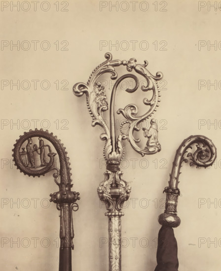 Three decorated episcopal staffs, displayed at an exhibition of religious objects from the Middle Ages and Renaissance in 1864 in Mechelen, photograph, page, number: '3 - 1 - 2', photographer: Joseph Maes, Mechelen, 1864, paper, albumen print, height 221 mm x width 184 mm, (liturgical) equipment of church, Period, Three ornate metal staffs with decorative crook tops are visible against a plain background, each featuring intricate scrollwork and sculpted figures; the central staff has a large, elaborate curved top with beaded edges, floral motifs, and small sculpted human figures, leading down to a detailed shaft with engraved patterns and a carved base; the staff on the left shows a coiled crook with a round framed scene of two standing figures and a textured shaft with decorative fittings; the staff on the right displays a narrower curled top with floral scrolls, a wrapped cloth hanging below an ornate collar, and a patterned handle.