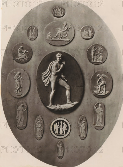 Medallion and fourteen cameos, photograph, page, photographer: Joseph Cundall, c. 1868 - in or before 1873, paper, carbon print, height 361 mm x width 270 mm, sculpture (relief, sculpture), carving, applied arts, An oval plaque with a central raised oval relief depicting a partially draped male figure standing with one foot on a rocky base and a cloak over his shoulder, surrounded by multiple smaller oval and circular relief medallions showing various classical-style figures in seated, standing, and interacting poses, all in a monochrome stone or plaster finish against a smooth background.
