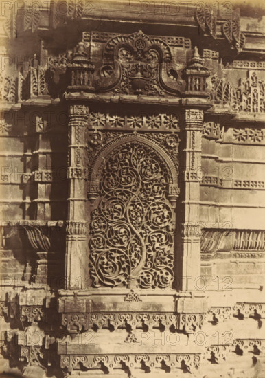 The Shápoor Mosque.- Niche from the base of the Northern Minaret, Decorations on a base of a minaret of the Shapoor Mosque in Ahmedabad, depicting a tree, photograph, page, number: '107.', photographer: Thomas Biggs, Ahmedabad (city), c. 1856 - in or before 1866, Gujarat, paper, albumen print, height 186 mm x width 136 mm, ornament derived from plant forms, temple, shrine, Islam, Mohammedanism, base (, column, pillar), A close view of an ornate stone wall section featuring a tall arched window with an intricate carved grille of intertwining vine and leaf patterns, flanked by decorated stone pilasters and framed by a carved arch with floral and scroll motifs; above the arch is a carved panel with symmetrical scrollwork and ornamental finials, and the surrounding masonry shows layered cornices, patterned friezes, and repeated floral and geometric reliefs with areas of shadow and weathering.