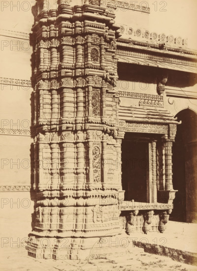 The Queen's Mosque in Mirzapoor.- Base of the Northern Minaret, Base of a minaret of the Rani Rupamati Mosque in Ahmedabad, photograph, page, number: '29.', inscription: 'I', photographer: Thomas Biggs, Ahmedabad (city), c. 1856 - in or before 1866, Thomas Biggs, photographer, Gujarat, paper, albumen print, height 189 mm x width 136 mm, temple, shrine, Islam, Mohammedanism, column, pillar, architecture, Rani Rupamati Mosque, A close view of a carved stone architectural facade showing a tall ornate pillar and an adjoining entrance section with decorative columns and a recessed dark doorway; the pillar surface is covered in layered horizontal bands and rectangular panels with intricate relief patterns and lattice-like circular motifs, and the entrance lintel and surrounding frieze display repeated ornamental borders and carved floral and geometric details; the lower part of the structure rests on a stepped base with rounded mouldings and bracket-like supports beneath the projecting platf...