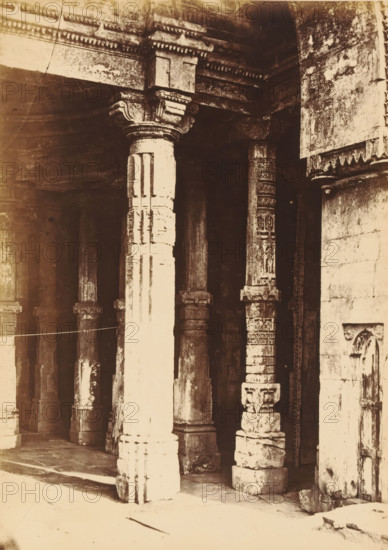 Ahmed Shah's Mosque.- Hindoo Pillars, Pillars of the Ahmed Shah's Mosque in Ahmedabad, photograph, page, number: '3.', photographer: Thomas Biggs, Ahmedabad (city), c. 1856 - in or before 1866, Gujarat, paper, albumen print, height 188 mm x width 136 mm, temple, shrine, Islam, Mohammedanism, column, pillar, architecture, Ahmed Shah Mosque, A sepia-toned photograph showing a row of carved stone columns and pillars forming a colonnaded interior, with one prominent lighter-colored column in the foreground featuring fluted vertical grooves and a detailed capital, adjacent to darker, weathered columns that display ornate carved patterns and decorative bands; the ceiling above the columns has layered cornices with geometric dentil-like molding, and the stone floor and wall surfaces show visible aging and mottled texture, with shadowed recesses between the pillars and fragments of carved stonework and a partially visible doorway at the right edge.
