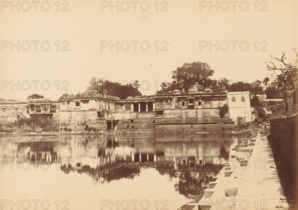 View of Sarkhej Roza near Ahmedabad, Sirkhej.- View from the South-east, photograph, page, number: '46.', inscription: 'N2', photographer: Thomas Biggs, Sarkhej Roza, c. 1856 - in or before 1866, Gujarat, paper, albumen print, height 136 mm x width 188 mm, grave, tomb, A sepia-toned photograph of a waterfront scene showing a stone building complex with domed roofs and arched openings set on stepped terraces leading down to calm water, trees rising behind the structures, a stone walkway with rectangular steps running along the right edge beside the water, and reflections of the buildings and trees visible on the water surface.