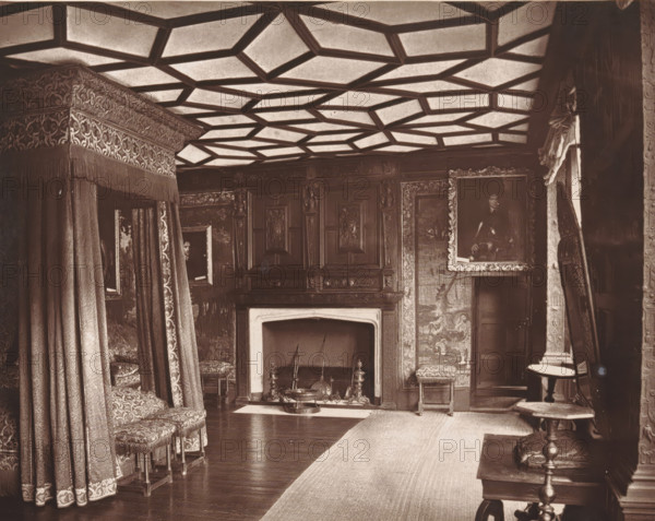 Interior of a bedroom at Knole House, The spangled bed-room, Knole, page, photomechanical print, photographer: Stephen Thompson, Knole House, c. 1866 - in or before 1876, printmaker, Kent, paper, Woodburytype, height 147 mm x width 185 mm, bedroom, interior of the house, A richly furnished interior with a patterned coffered ceiling, ornate draperies forming a canopy over a bed area at left, upholstered stools with decorative fabric and fringe, a carved wooden fireplace surround with a mantel, and a hearth containing fireplace tools andirons and a low decorative brazier; carved wooden paneling and framed paintings hang on the walls, one large portrait in an ornate frame at right, a tapestry or wall covering with figures beside the fireplace, a narrow rug running along a polished wooden floor, a small round table with a turned pedestal and an upholstered footstool nearby, and heavy curtains and a carved doorway visible at the far right.