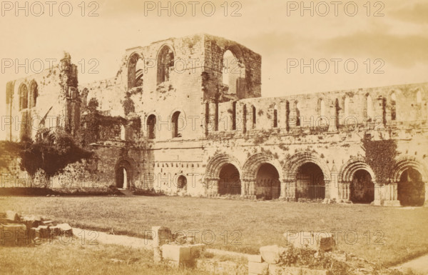 View of the ruins of Furness Abbey, Furness Abbey, from the south-west, photograph, page, photographer: Garnett & Sproat, c. 1857 - c. 1867, Cumbria, paper, albumen print, height 106 mm x width 166 mm, ruin of church, monastery, A sepia-toned photograph of a large stone ruin with tall arched openings and a series of smaller arched arcades along its façade, showing weathered masonry and patches of climbing vegetation on the walls, with a grassy foreground scattered with stone blocks and remnants, and parts of the structure featuring large window openings and recessed doorways.