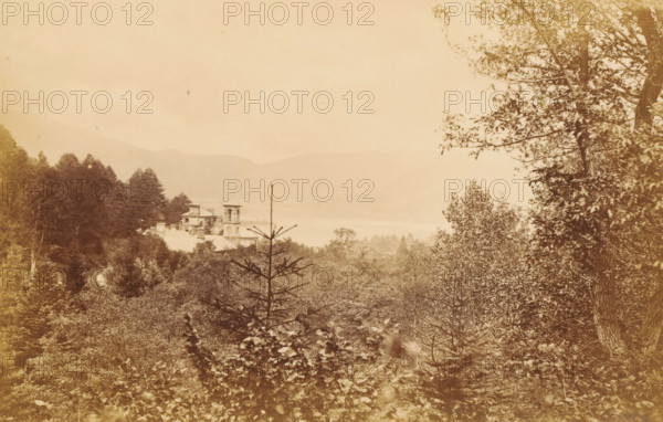 Ullswater, from Grisedale, View of Ullswater in the Lake District, photograph, page, photographer: Garnett & Sproat, Lake District, c. 1857 - c. 1867, paper, albumen print, height 105 mm x width 165 mm, lake, forest, wood, mountains, A sepia-toned landscape showing dense trees and foliage in the foreground and middle ground with a narrow conifer protruding centrally, a multi-tiered building with a square tower partially visible among the trees to the left of center, a calm body of water behind the building, distant hazy hills across the horizon, and leafy branches framing the right edge of the image.