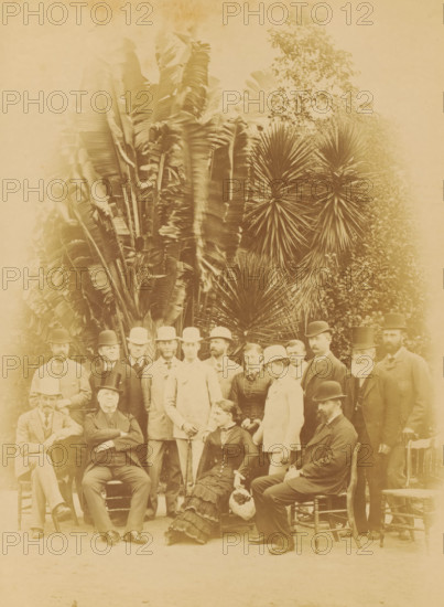 Group portrait of Arthur Kennedy and his family, Prince George of Wales and Prince Albert Victor of Wales in Brisbane, photograph, page, Brisbane (Australia), c. 1880 - in or before 1881, photographer, Queensland, paper, albumen print, height 226 mm x width 168 mm, Person, Albert Victor (Prince of the United Kingdom), George V (King of the United Kingdom), Arthur Kennedy, colonial governor, anonymous historical persons portrayed in a group, in a group-portrait, prince, crown prince, A sepia-toned group portrait shows a mix of men and women posed outdoors in front of large spiky and broad-leafed plants; the men wear suits and various styles of hats including bowler and top hats, and several have facial hair, while the women wear long dresses with high collars and hats; some people are seated on wooden chairs and others stand closely behind them, with one seated woman holding a round woven basket, and a few men hold walking sticks.
