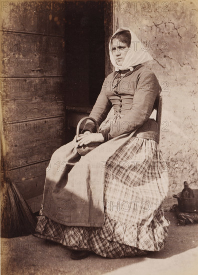 Portrait of an unknown woman on a chair, photograph, page, Italy, c. 1850 - in or before 1860, photographer, paper, albumen print, height 135 mm x width 96 mm, adult woman, historical persons not known by name - BB - woman, A seated woman wearing a headscarf and layered dress sits near a wooden door and a cracked plaster wall, holding a small rounded basket in her lap with both hands, her gaze directed slightly away from the camera, with a broom leaning against the door and a metal pot on the ground beside her.