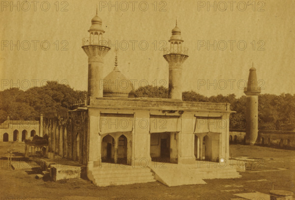 View of the Karbala Talkatora in Lucknow, Kurballa Talkatora, photograph, page, number: '50', photographer: Darogha Ubbas Alli, Lucknow, c. 1864 - in or before 1874, Oudh, paper, albumen print, height 105 mm x width 154 mm, temple, shrine, Islam, Mohammedanism, A sepia-toned photograph of a building complex with architectural elements including a domed roof and several cylindrical towers topped with ornate finials and balcony-like platforms; the foreground structure has a colonnaded façade with arched openings and a short flight of steps leading up to it, and portions of the walls show weathering and dark streaks; behind and beside the main building are dense clusters of trees and additional arched structures extending along one side; the ground in front shows patches of grass, stone slabs, and low rectangular stone blocks scattered around.
