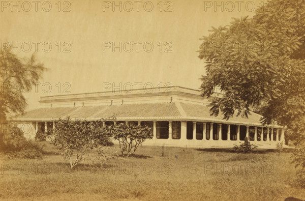 Darus-shafa, View of a hospital, Dar-us Shafa, in Lucknow, photograph, page, number: '8', photographer: Darogha Ubbas Alli, Lucknow, c. 1864 - in or before 1874, Oudh, paper, albumen print, height 105 mm x width 155 mm, hospital, A sepia-toned photograph shows a single-story building with a long tiled roof and a covered veranda supported by columns, dark window openings along the facade, and decorative cornice detail along the roofline; the building is set behind a grassy foreground with small leafy shrubs and trees, and larger trees frame the scene on both sides.