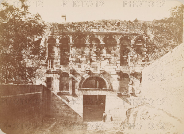 Porta Aurea of the Palace of Diocletian in Split, Porta Aurea, Spalato und seine Alterthümer, photograph, page, photographer: Franz Thiard de Laforest, Split, c. 1868 - in or before 1878, Yugoslavia, Splitsko-Dalmatinska Županija, paper, albumen print, height 100 mm x width 137 mm, city-gate, Diocletian's Palace, A sepia-toned photograph showing the façade of a stone building with a series of arched openings along its upper portion and a large arched doorway below, flanked by rectangular stone blocks and architectural detailing. Trees and foliage frame the left and right edges of the image, partially obscuring the upper wall on the left. A slope or ramp leads up to the entrance area, casting diagonal shadows across the lower façade. A person stands near the base of the entrance with a small dark animal nearby. The stone surfaces show weathering and irregular texture across the entire structure.