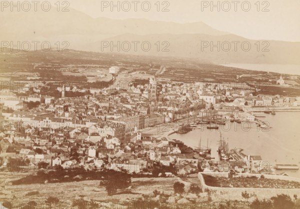 Spalato, View of Split, Spalato and seine Alterthümer, photograph, page, photographer: Franz Thiard de Laforest, Splitsko-Dalmatinska Županija, c. 1868 - in or before 1878, Yugoslavia, paper, albumen print, height 103 mm x width 148 mm, city-view in general, 'veduta', Split, A sepia-toned panoramic view shows a coastal town with densely packed buildings and narrow streets extending toward a waterfront where several sailing ships are moored along curved quays and piers; among the buildings are taller structures with towers and steeples rising above lower rooftops, and a long row of multi-story facades faces the harbor; beyond the town there are terraced fields and low stone walls stretching across a gently rolling landscape that leads to distant layered hills and mountains under a pale sky.