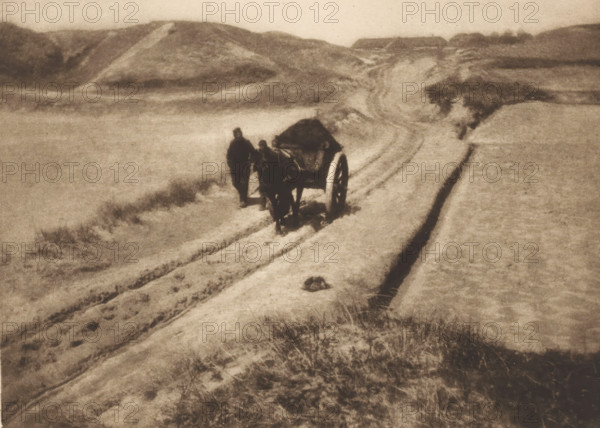 View of two unknown men with a cart on a path through the dunes, The road through the dunes, page, photomechanical print, photographer: James Craig Annan, cliché maker: Meisenbach, Riffarth & Cie., Berlin, publisher: Wilhelm Knapp Verlag, Halle an der Saale, c. 1897 - in or before 1902, Saxony-Anhalt, paper, photoaquatint, height 146 mm x width 203 mm, dunes (sea not visible), in the dunes, road, path, hand-cart, A grainy monochrome scene shows a sandy landscape with undulating dunes and a winding track cutting through the sand. Near the center a covered cart with large wheels stands on the track, accompanied by two upright figures close by. Tire or wheel ruts run along the path leading toward distant low hills. Sparse tufts of grass and scattered vegetation appear in the foreground and along the edges of the track. The overall tones are muted and sepia-like, with soft contrasts and a hazy sky above the dunes.