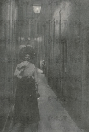 Gasse in Venedig, Portrait of an unknown woman with fan and hat in an alley in Venice, page, photomechanical print, photographer: Alfred Kirstein, Venice, cliché maker: Meisenbach, Riffarth & Cie. (possibly), Berlin, publisher: Wilhelm Knapp Verlag, Halle an der Saale, c. 1897 - in or before 1902, Saxony-Anhalt, paper, autotypie, height 212 mm x width 145 mm, Print seven pages after page 102., adult woman, anonymous historical person depicted, A narrow, dimly lit alley with tall walls on either side and a light fixture mounted high above; a woman stands in the foreground wearing a dark skirt and a light blouse, holding a folded hand fan partly in front of her face and carrying a small handbag, with another figure behind her partly visible and a long, pale cloth or garment trailing along the ground down the alley.