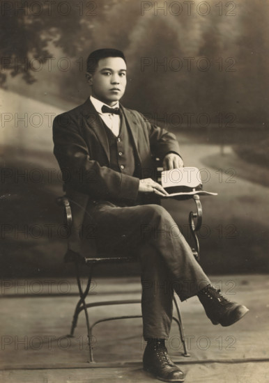 Portrait of an unknown Asian man, photograph, address: '????????????, ??????????? No 37, ???????? No 544', inscription: 'Huronán, Ban-uiu-ho ?', photographer: Chvunchva, Vladivostok, c. 1910 - c. 1930, Tsjvoenkhva, paper, pasteboard, baryta paper, gelatin silver print, developing gelatin silver print, height 144 mm x width 103 mm, height 170 mm x width 105 mm, adult man, anonymous historical person depicted, A seated young man in a dark three-piece suit and bow tie faces the camera with a neutral expression, one arm resting on the back of a metal chair and the other hand holding a light-colored hat placed on his knee, his legs crossed to show dark lace-up boots; the background is softly blurred with indistinct foliage and a path, and the image has a sepia-toned, vintage appearance.