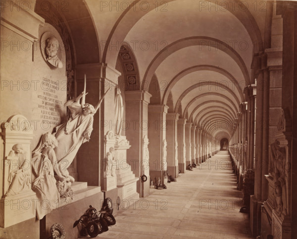 Genova Camposanto Galleria, Sculpture gallery with funeral wreaths at the Staglieno cemetery in Genoa, photograph, Part of French artist's album., Campo Santo (Genoa), c. 1880 - c. 1900, photographer, Genoa, paper, albumen print, height 218 mm x width 261 mm, gallery of sculptures, cemetery, park-like burying ground, A long covered corridor with repeating rounded arches and columns stretching into the distance, flanked by sculpted wall monuments and statues along one side, including a reclining angel figure and other carved human figures, a circular portrait relief high on the wall, decorative floral ceiling panels visible in some arch intrados, wreaths and floral arrangements placed at the bases of several monuments, a tiled or stone floor extending toward a small distant altar or niche, and a cloak-draped statue among the memorials.