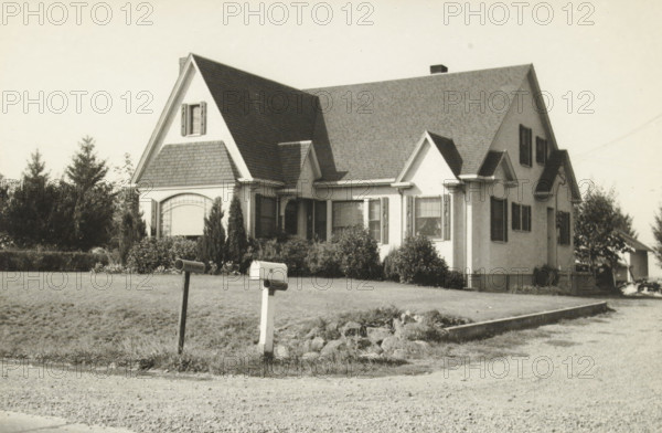 A country house in the west. Note the newspapers and mailboxes, Unknown location in the western United States: a country house with mailboxes by the road, photograph, title: 'A COUNTRY HOUSE IN THE WEST, NOTE THE NEWSPAPERS AND MAILBOXES', photographer: attributed to Wouter Cool, United States of America, 1936, North America, Attributed to, pasteboard, paper, gelatin silver print, gelatin silver developing print, height 151 mm x width 229 mm, height 315 mm x width 285 mm, country-house, A single-story house with a steep gabled roof and multiple dormers sits behind a front lawn, flanked by tall shrubs and small trees; the house has shuttered windows, a small covered porch area, and decorative siding on a front gable section, while two mailboxes on short posts stand near the edge of the lawn by a gravel road and a low stone-bordered strip separates the yard from the driveway.