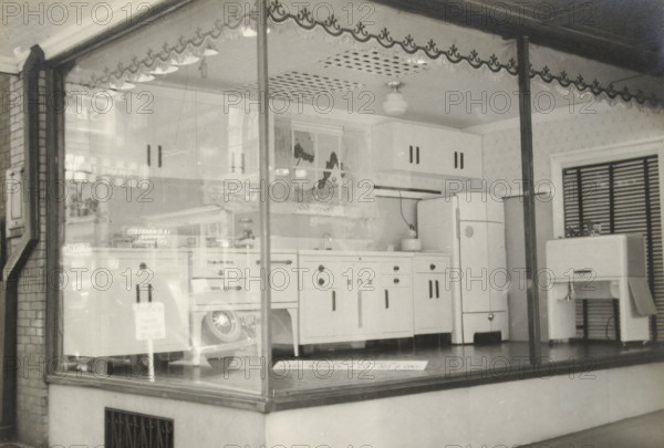 A Shop Window Displaying an Electric Kitchen, photograph, title: 'WINDOW WITH ELECTRIC KITCHEN EQUIPMENT', photographer: attributed to Wouter Cool, United States of America, 1936, North America, Attributed to, pasteboard, paper, gelatin silver print, developing gelatin silver print, height 154 mm x width 224 mm, height 315 mm x width 285 mm, kitchen, shop window, show window, A black and white photograph shows a storefront window display of a kitchen scene with a freestanding refrigerator, a sink cabinet with drawers and doors, an oven with control knobs, and an under-sink cabinet; a small appliance sits on the countertop beside the sink and a separate small table or cabinet stands to the right with an object on top; patterned wallpaper covers the rear wall and a louvered window or shutter is visible on the right; decorative scalloped trim runs along the top inside the window and a ceiling light fixture hangs above; the display is framed by large glass panes with metal supports and a b...