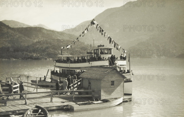 The Little Boat JD Ross, A boat on a lake at an unknown location in the United States, photograph, title: 'THE LITTLE BOAT, JD ROSS', photographer: attributed to Wouter Cool, United States of America, 1936, North America, Attributed to, pasteboard, paper, gelatin silver print, height 145 mm x width 220 mm, height 285 mm x width 315 mm, lake, traffic on the water, A dock extends into a calm body of water with a wooden walkway and railings leading to a larger dock structure where people stand and board a two-deck passenger boat decorated with a string of triangular flags draped from a mast to the bow. The lower deck of the boat is partially enclosed while the upper deck has railings and a small enclosed pilothouse with windows; silhouettes of people are visible on both decks and on the dock. A small shed with a slanted roof sits on the dock adjacent to the boat, and a rowboat is moored alongside the dock in the foreground. In the background, layered hills and mountains rise from the wate...