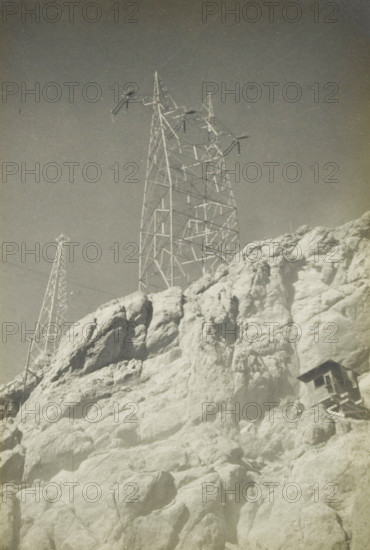 280 kv masts, Electricity pylons at an unknown location in the United States, photograph, title: '280 kV MASTEN', photographer: attributed to Wouter Cool, United States of America, 1936, North America, Attributed to, pasteboard, paper, gelatin silver print, developing gelatin silver print, height 228 mm x width 152 mm, height 315 mm x width 215 mm, pylons, power lines, A tall metal lattice tower with crossarms and insulators rises above a rocky cliff, alongside a smaller lattice tower visible further back, both set against a clear sky; the cliff shows rough, light-colored rock formations with cracks and shadows, and a small wooden structure is perched on the lower right face of the rocks.