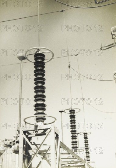 Earthing switches in electrical lines, Earthing switches of the 280 kV line, photograph, title: 'EARTHING SWITCHES, OF THE 280 kV LINE.', photographer: attributed to Wouter Cool, United States of America, 1936, North America, Attributed to, pasteboard, paper, gelatin silver print, developing gelatin silver print, height 226 mm x width 158 mm, height 315 mm x width 215 mm, power station, The image shows a series of tall insulator stacks with ribbed, disc-like sections mounted on metal frameworks and topped by circular metal rings, with thin suspension wires and curved overhead cables stretching across a pale sky, and portions of structural beams and electrical hardware visible at the bottom and edges.