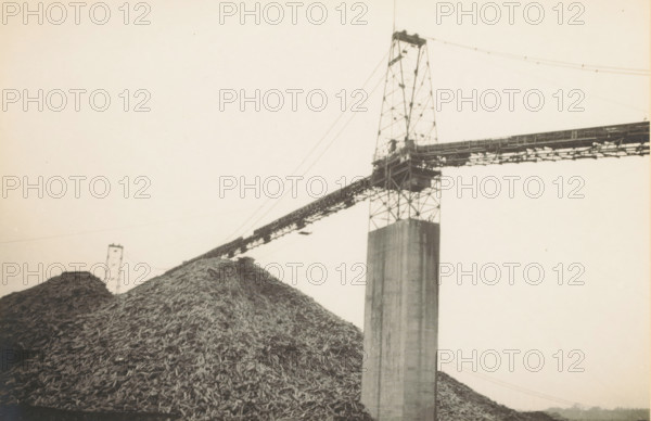 Wood pile for Grand Mere paper factory, Wood pile at paper factory in Grand Mère, near Montréal, Canada, photograph, title: 'WOOD STACK FOR, GRAND MERE PAPER FACTORY', photographer: attributed to Wouter Cool, United States of America, 1936, North America, Attributed to, pasteboard, paper, gelatin silver print, developing gelatin silver print, height 152 mm x width 224 mm, height 315 mm x width 286 mm, industrial structures, A large pile of irregular wooden pieces fills the lower left and center foreground, with a tall concrete column rising from the pile toward the right. A steel lattice tower sits atop the column and supports a long horizontal conveyor structure that extends across the image, with cables running from the tower to another distant lattice support behind the pile. The scene is monochrome with a pale sky background and fine grain visible in the image.