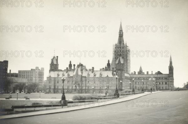 Parliament Buildings, Ottawa, Canada, Government Building Ottawa, photograph, title: 'GOVERNMENT BUILDING, TE OTTAWA CANADA.', photographer: attributed to Wouter Cool, United States of America, 1936, North America, Attributed to, pasteboard, paper, gelatin silver print, developing gelatin silver print, height 153 mm x width 226 mm, height 315 mm x width 272 mm, government, the State, 'Politica', 'Auttorità o Potestà' (Ripa), A wide view of a large stone building with multiple steep roofs and pointed towers, one tall central tower surrounded by scaffolding, rows of arched windows and decorative stonework along its facade, lamp posts lining a paved walkway in front, a grassy area with low stone walls and benches between the building and the road, a few distant pedestrians on the sidewalk, and additional multi-story buildings visible beyond and beside the main structure under a pale sky.