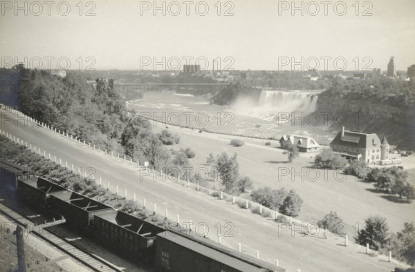 Niagara Falls, United States, Canada, Amerikasche Vrouwen, photograph, title: 'NIAGARA FALLS, AMERIKAANSCHE VALLEN.', photographer: attributed to Wouter Cool, United States of America, 1936, North America, Attributed to, pasteboard, paper, gelatin silver print, developing gelatin silver print, height 154 mm x width 229 mm, height 315 mm x width 285 mm, waterfall, A wide black-and-white view shows a river with a large waterfall and mist rising where the water drops, a bridge crossing the river in the distance, and a shoreline with trees and grassy open areas. In the foreground a sloped paved road runs diagonally with regularly spaced short posts along its edge, and a set of parallel railroad tracks with open freight cars sits at the bottom edge. Two buildings with pitched roofs and a larger multi-story building with a turret stand near the riverbank, with scattered trees and small shrubs around them. The far skyline includes low industrial and urban structures beyond the river and a smo...