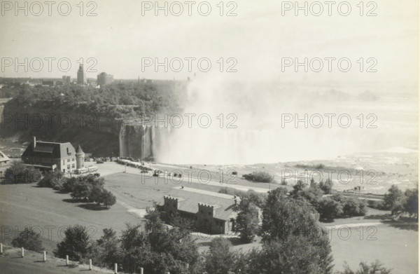 Niagara Falls (Horse Shoe), United States, Canada, Niagara Falls Horse shoe, photograph, title: 'NIAGARA FALLS, HORSE SHOE.', photographer: attributed to Wouter Cool, United States of America, 1936, North America, Attributed to, pasteboard, paper, gelatin silver print, developing gelatin silver print, height 154 mm x width 225 mm, height 285 mm x width 315 mm, waterfall, A wide aerial view shows a large waterfall with a heavy mist rising and spreading across the scene, cliffs and cascading water visible where the falls meet the river below. In the foreground there are grassy lawns dotted with trees, a roadway lined with parked cars and a few buses, and a substantial stone building with a steep roof and a small round turret near its corner. Nearby are smaller castle-like stone structures with battlemented tops, and a low stone wall edging the foreground. Beyond the falls a tree-covered ridge and a distant city skyline with several tall buildings and towers are visible under a pale sky.