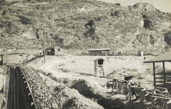 Grand Coulee dam gravel supply to the concrete mixer, Grand Coulee Dam, Columbia River, United States: gravel supply, photograph, title: 'GRAND COULEE DAM, GRAVEL SUPPLY TO THE CONCRETE MILL', photographer: attributed to Wouter Cool, United States of America, 1936, North America, Attributed to, pasteboard, paper, gelatin silver print, developing gelatin silver print, height 150 mm x width 229 mm, height 315 mm x width 330 mm, dam (in canal), A sepia-toned landscape showing a rocky hillside with sparse vegetation and exposed rock faces, a narrow dirt road winding along the midground, small wooden buildings and sheds scattered near the road, utility poles and power lines crossing the scene, a raised wooden trestle carrying a conveyor or chute filled with rocks extending from the foreground toward the left, an open wooden tower structure standing near the road, assorted wooden scaffolding and crates in the lower right, and a hillside sign with block letters near the top edge.