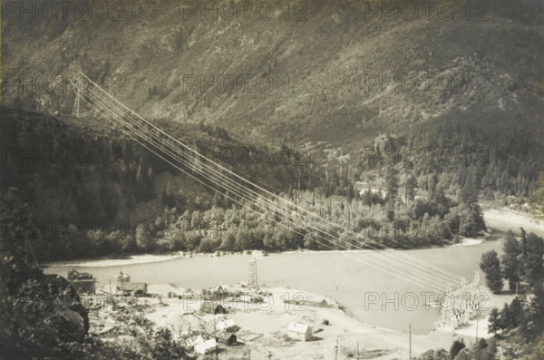 Line: Diabolo dam power station gorge, Electricity connection between Gorge power station and the Diablo Dam, Washington state, United States, photograph, title: 'LINE : DIABOLO DAM-, CENTRAL GORGE', photographer: attributed to Wouter Cool, United States of America, 1936, North America, Attributed to, pasteboard, paper, gelatin silver print, developing gelatin silver print, height 153 mm x width 227 mm, height 315 mm x width 285 mm, pylons, power lines, A grayscale landscape showing a river bordered by a treed shoreline and a forested hillside rising behind it, with electrical transmission towers and multiple power lines crossing diagonally from the upper left toward the lower right over the river, a small cluster of buildings and scattered structures near the near bank, a gravelly or sandy area in the foreground with small sheds and debris, and a boat or floating structure moored along the far bank.