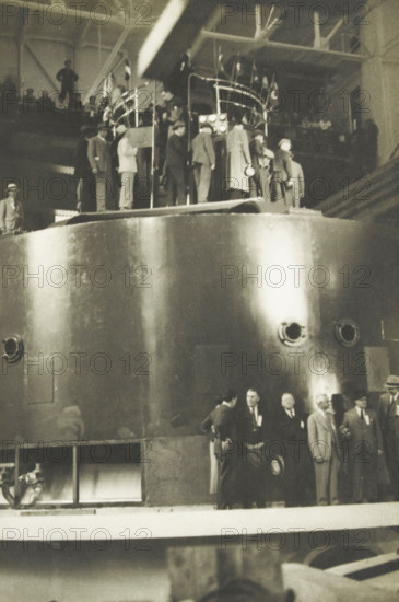 Water turbo of the Diablo Dam, Washington state, United States, Large water turbo 90, 000 kva central diabolo dam, photograph, title: 'LARGE WATER TURBORE 90, 000 kVA, CENTRAL DIABOLO DAM.', photographer: attributed to Wouter Cool, United States of America, 1936, North America, Attributed to, pasteboard, paper, gelatin silver print, developing gelatin silver print, height 227 mm x width 151 mm, height 315 mm x width 330 mm, dam (in canal), A large metallic cylindrical structure fills most of the image, its curved surface reflecting light and showing round openings and a small rectangular panel. Along the top edge of the cylinder a group of people stand on a platform, some wearing coats and hats and some clustered near a railing with vertical posts and lights. More people in suits and hats are visible at a lower level near the base of the cylinder, some standing and some seated, with a few facing each other in conversation. In the upper background an elevated walkway or balcony with str...