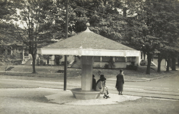 Tram stop in Seattle, Washington state, United States, Tram house in Seattle, photograph, title: 'TRAM HOUSE, IN SEATTLE', photographer: attributed to Wouter Cool, United States of America, 1936, North America, Attributed to, pasteboard, paper, gelatin silver print, developing gelatin silver print, height 142 mm x width 223 mm, height 315 mm x width 285 mm, tram, A grayscale photograph shows a sheltered street-side structure with a broad, shingled, pyramidal roof topped by a small finial, supported by a central column with a bench-like base. Several people are near and under the shelter; one person stands facing away from the camera while others sit on the bench, wearing long coats and hats. Behind the shelter is a tree-lined residential street with houses, porches, steps, and large trees with dense foliage. A lamppost stands next to the shelter and the foreground includes a paved area and streetcar tracks running parallel to the curb.