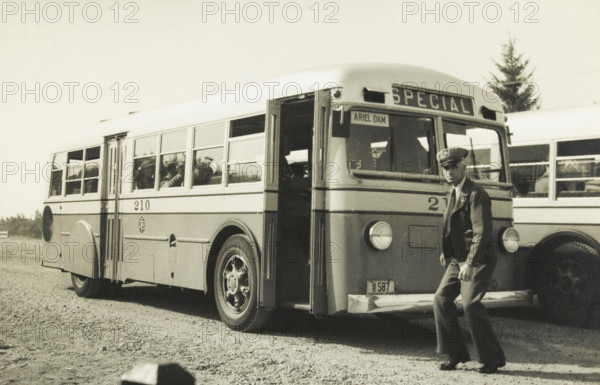 The newest buses, pneumatic brakes and gears, rear engine, Ariel Dam, Washington state, United States: bus, photograph, title: 'THE NEWEST BUSES, PNEUMATIC, BRAKES AND GEARS, REAR ENGINE.', photographer: attributed to Wouter Cool, United States of America, 1936, North America, Attributed to, pasteboard, paper, gelatin silver print, developing gelatin silver print, height 154 mm x width 228 mm, height 315 mm x width 285 mm, coach, A vintage bus parked on a gravel surface with its front door open and a man in a uniformed jacket and cap standing near the front bumper, the bus showing a row of windows along its side with passengers visible through them, round headlights on the front, large wheels with visible hubcaps, another similar bus partially visible behind it, and a tree in the background under a clear sky.