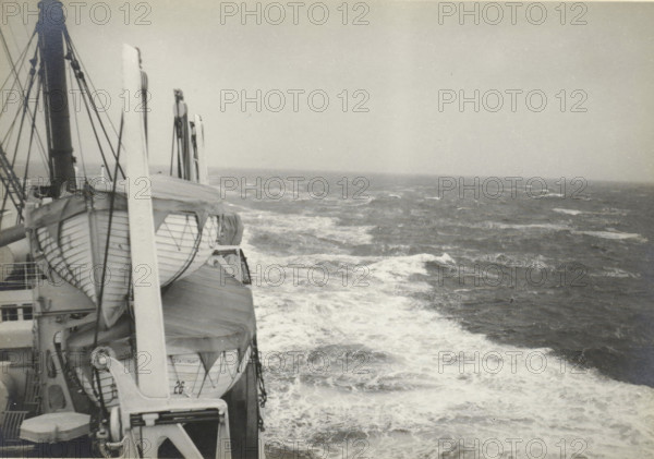 During the crossing from France to the United States, Atlantic Ocean, Storm from behind, photograph, title: 'STORM FROM BEHIND.', photographer: attributed to Wouter Cool, United States of America, 1936, North America, Attributed to, pasteboard, paper, gelatin silver print, height 154 mm x width 222 mm, height 315 mm x width 286 mm, steamship, motorship, The image shows a ship's deck area with lifeboats secured in cradles along the rail, rigging lines and davits rising upward, and canvas covers over parts of the boats; to the right, the open sea stretches to the horizon with waves and white foam trailing alongside the vessel under a mostly pale sky.