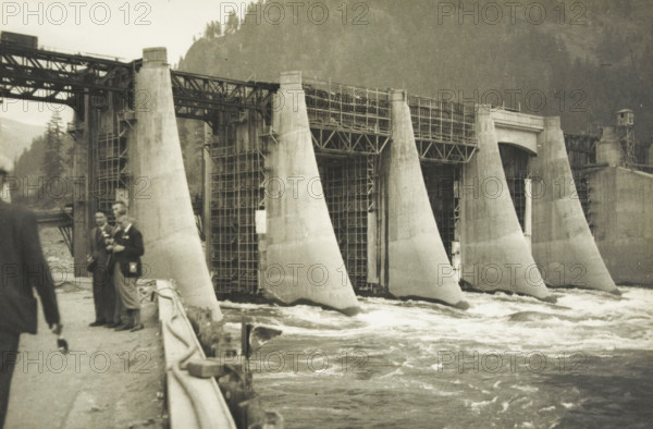 Bonnerville dam, Portland Oregon, Portland, Oregon, United States, photograph, title: 'BONNERVILLE DAM, PORTLAND-OREGON', photographer: attributed to Wouter Cool, United States of America, 1936, North America, Attributed to, pasteboard, paper, gelatin silver print, developing gelatin silver print, height 153 mm x width 228 mm, height 315 mm x width 330 mm, dam (in canal), A sepia-toned scene of a large concrete dam with several tapered piers rising from turbulent water, scaffolding and metal framework visible between the piers and along the top, a walkway or road at the near left edge with a railing draped with fabric, and a few people standing on the walkway wearing long coats and hats, with a mountainous, tree-covered slope in the background.