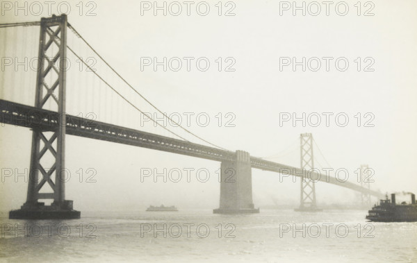 Oakland Bridge, San Francisco, photograph, title: 'OAKLAND BRIDGE, SAN FRANCISCO', photographer: attributed to Wouter Cool, United States of America, 1936, North America, Attributed to, pasteboard, paper, gelatin silver print, developing gelatin silver print, height 150 mm x width 229 mm, height 315 mm x width 285 mm, bridge, A wide view of a suspension bridge spanning across a body of water, shown in a monochrome tone, with tall lattice bridge towers and diagonal suspension cables extending from the left foreground toward the right background, a central bridge pier rising from the water beneath the roadway, a small ship visible beneath the bridge near the center, and a ferry or boat at the right edge partially entering the frame, with the water surface shimmering and the sky appearing light and hazy.