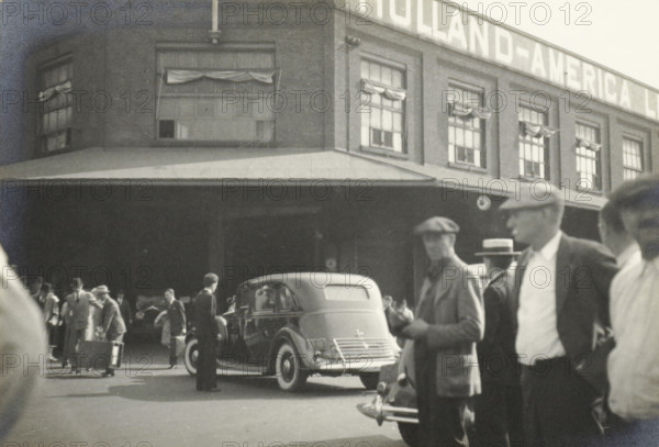 The Pier of the Holland America Line in Hoboken, New York, photograph, title: 'ENTANG PIER, TE HOBOKEN.', photographer: attributed to Wouter Cool, United States of America, 1936, North America, Attributed to, pasteboard, paper, gelatin silver print, developing gelatin silver print, height 158 mm x width 230 mm, height 315 mm x width 286 mm, pier, quay, wharf, A street scene in front of a brick building with multiple windows, some with fabric draped above them and a corrugated awning along the façade, shows a parked vintage car with rounded rear and visible bumper and spare tire, and a crowd of people standing and walking near the vehicle, many wearing hats and coats and carrying suitcases or bags.