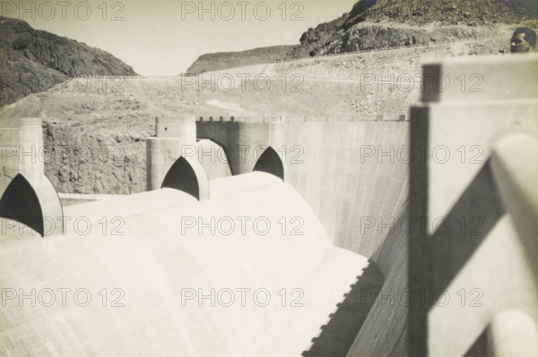 Boulder Dam, Arizona, Nevada, United States, Spillwaij Boulderdam, photograph, title: 'SPILLWAIJ BOULDERDAM', photographer: attributed to Wouter Cool, United States of America, 1936, North America, Attributed to, pasteboard, paper, gelatin silver print, developing gelatin silver print, height 154 mm x width 227 mm, height 315 mm x width 330 mm, dam (in canal), A high concrete dam with curved spillway faces and three arched openings along its crest, light and shadow creating strong contrasts across the smooth concrete surfaces; rocky hills rise behind the dam with rough, textured slopes and a winding road along the ridge; a vertical concrete barrier casts a triangular shadow in the foreground and a partial head of a person appears near the top edge above the barrier.