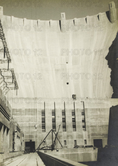 Boulder Dam, Arizona, Nevada, United States, Centrale Boulder dam, photograph, title: 'CENTRALE BOULDERDAM', photographer: attributed to Wouter Cool, United States of America, 1936, North America, Attributed to, pasteboard, paper, gelatin silver print, developing gelatin silver print, height 227 mm x width 158 mm, height 315 mm x width 330 mm, dam (in canal), A large concrete curved wall rises from the bottom to the top of the image, showing vertical texture and rectangular recesses along its upper edge, with a row of narrow vertical slits near the lower portion; a horizontal ledge and a darker rectangular opening sit near the base, and a small box-like structure is mounted above the ledge. In the foreground a flat roadway or walkway leads toward the base, bordered by a low concrete barrier on one side and an open channel on the other, with a small vehicle visible entering a shadowed opening. A metal crane-like framework with angled beams is positioned near the center, and a series of ...