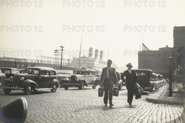 Arrival in New York: the SS Statendam, Hoboken pier, United States, SS Statendam on the pier in Hoboken, photograph, title: 'SS STATENDAM ON THE PIER, HOBOKEN.', photographer: attributed to Wouter Cool, United States of America, 1936, North America, Attributed to, pasteboard, paper, gelatin silver print, developing gelatin silver print, height 153 mm x width 228 mm, height 315 mm x width 286 mm, pier, quay, wharf, A black and white street scene showing a cobblestone road filled with parked vintage cars and taxis, with two men walking toward the camera carrying suitcases; one man wears a light-colored suit and tie while the other wears a dark suit and a hat. In the background a large ocean liner with multiple funnels is docked behind a fence and industrial structures, and buildings line the right side of the street with a lamppost and fence visible near the sidewalk.