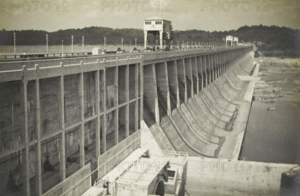 Conowingo Dam, near Perryville, Maryland, United States, Conowingodam near Perrijville, photograph, title: 'CONOWINGODAM NEAR, PERRIJVILLE.', photographer: attributed to Wouter Cool, United States of America, 1936, North America, Attributed to, pasteboard, paper, gelatin silver print, developing gelatin silver print, height 153 mm x width 227 mm, height 315 mm x width 330 mm, dam (in canal), A long concrete dam stretches diagonally across the image with a roadway along its top lined by slender poles and small structures, and a blocky control tower set on tall supports visible near the center; the dam face shows vertical supports and curved spillway sections descending toward a calm body of water at the right, while the background features a tree-covered shoreline and a hazy sky.