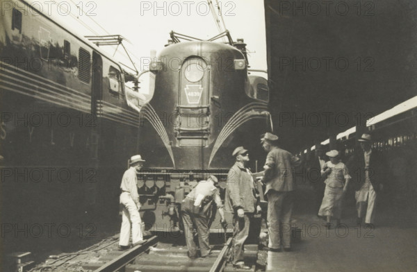 Electric locomotive of the Pensijlvania railroad, Electric locomotive of the Pennsylvania Rail Road, United States, photograph, title: 'ELECTRIC LOCOMOTIVE, OF THE PENSIJLVANIA RAILROAD.', photographer: attributed to Wouter Cool, United States of America, 1936, North America, Attributed to, pasteboard, paper, gelatin silver print, developing gelatin silver print, height 148 mm x width 220 mm, height 315 mm x width 285 mm, locomotive, A large streamlined train locomotive fills the center of the scene with curved metal surfaces and a prominent round headlight, flanked by parallel train cars on either side and overhead electrical wires. Several men stand and work on and around the front of the locomotive, wearing hats and work clothes, one leaning over near the coupling and another holding a tool while others converse. On the platform to the right a woman in a dress and hat walks while another man in a hat and long coat stands nearby carrying a bag. The image is monochrome and shows the p...