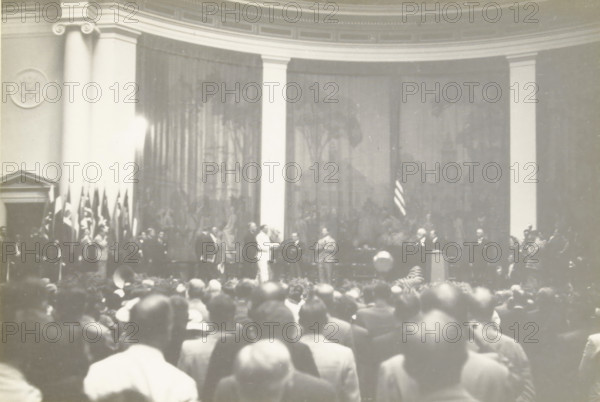 President Roosevelt enters the convention hall, Constitution Hall Washington, Speech by US President Franklin D. Roosevelt during the Third World Power Conference, Constitution Hall, Washington DC, United States, photograph, title: 'PRESIDENT ROOSEVELT ENTERS THE CONSCIENCE HALL, CONSTITUTION HALL WASHINGTON', photographer: attributed to Wouter Cool, United States of America, 1936-09-11, North America, Attributed to, pasteboard, paper, gelatin silver print, gelatin silver developing print, height 153 mm x width 228 mm, height 315 mm x width 285 mm, president, head of republic, A wide indoor scene showing a large audience in the foreground facing a raised stage area where a group of people stand and interact; tall columns frame a backdrop with a curtain and faint decorative patterns, and a display of vertical flags stands to one side while a single flag hangs near the center; several men in suits and one person in a light-colored outfit are positioned across the stage, with a podium and...