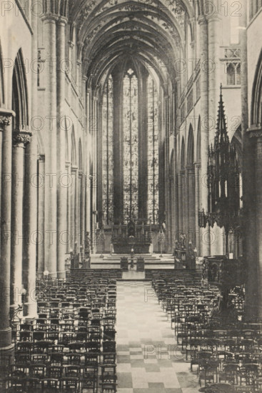 Huy. - La Collégiale, Interior of the Collegiate Church of Our Lady and Domitian of Huy, page, photomechanical print, photographer: attributed to Victor Gaillard, Huy, cliché maker: attributed to Emile Aubry, Brussels, c. 1885 - in or before 1890, Hoei, Attributed to, paper, collotype, photoprint, height 158 mm x width 105 mm, interior of church, A long interior view of a vaulted stone building with tall ribbed arches and a high stained glass window at the far end, a central aisle with a checkered floor leading toward an altar area, rows of wooden chairs arranged on either side of the aisle, tall cylindrical columns with carved capitals lining both sides, ornate carved stone and metalwork visible along the right side including a dark, pinnacled structure, and soft light filtering through the large vertical window panels.