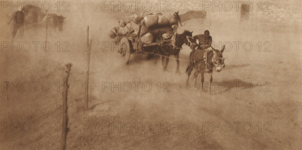 Path with cart drawn by donkeys, page, photomechanical print, photographer: Donald Mennie, Beijing, cliché maker: anonymous, c. 1915 - in or before 1920, paper, rotogravure, height 152 mm x width 291 mm, road, path, animal traction, animal-drawn vehicle (carriage etc.), China, A sepia-toned scene shows a dusty open area with a loaded wooden cart pulled by two donkeys and guided by a person sitting on the cart, another donkey in front of the cart wearing a bridle and harness, and scattered thin wooden posts rising from the ground; the cart is piled with large sacks and bundles, and the dust creates a hazy atmosphere that softens edges and mutes background details.