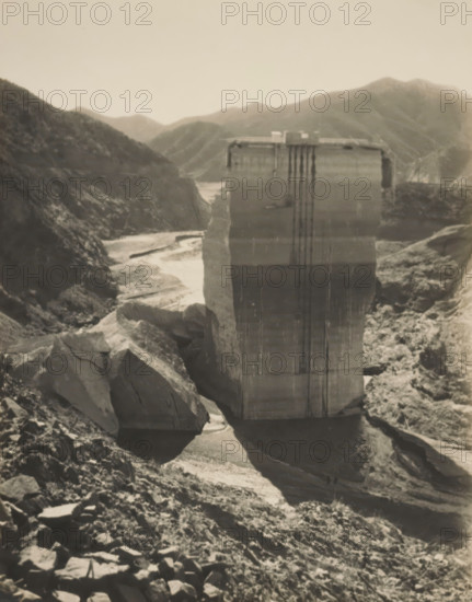 Saint Francis Dam, album page, photograph, annotation: 'Sint Francesdam restant (300.000.000 lit water)', annotation: 'San Juan Capistrano', annotation: '300.000.000 L. water March 1928 St. Frances Dam', California, 1928-03, photographer, North America, paper, gelatin silver print, height 254 mm x width 200 mm, dammed-up lake, Los Angeles (county), A large, weathered concrete pillar stands in the center of a rocky canyon, its surface marked by vertical streaks and horizontal layers, with a flat top that has small structures and railings. Steep, jagged rock faces and boulders surround the pillar on both sides, forming a narrow, winding dry riverbed that curves away toward distant, rolling hills. The foreground shows a slope strewn with loose rocks and rubble, casting shadows across uneven ground, while the distant background consists of layered mountain ridges under a pale sky.